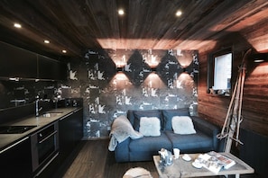 Une cuisine moderne avec un revêtement mural foncé, un four encastré et un évier. On y trouve un canapé bleu foncé avec des coussins blancs et une couverture, une table basse en bois avec des magazines et une tasse, et un mur en bois avec une fenêtre.