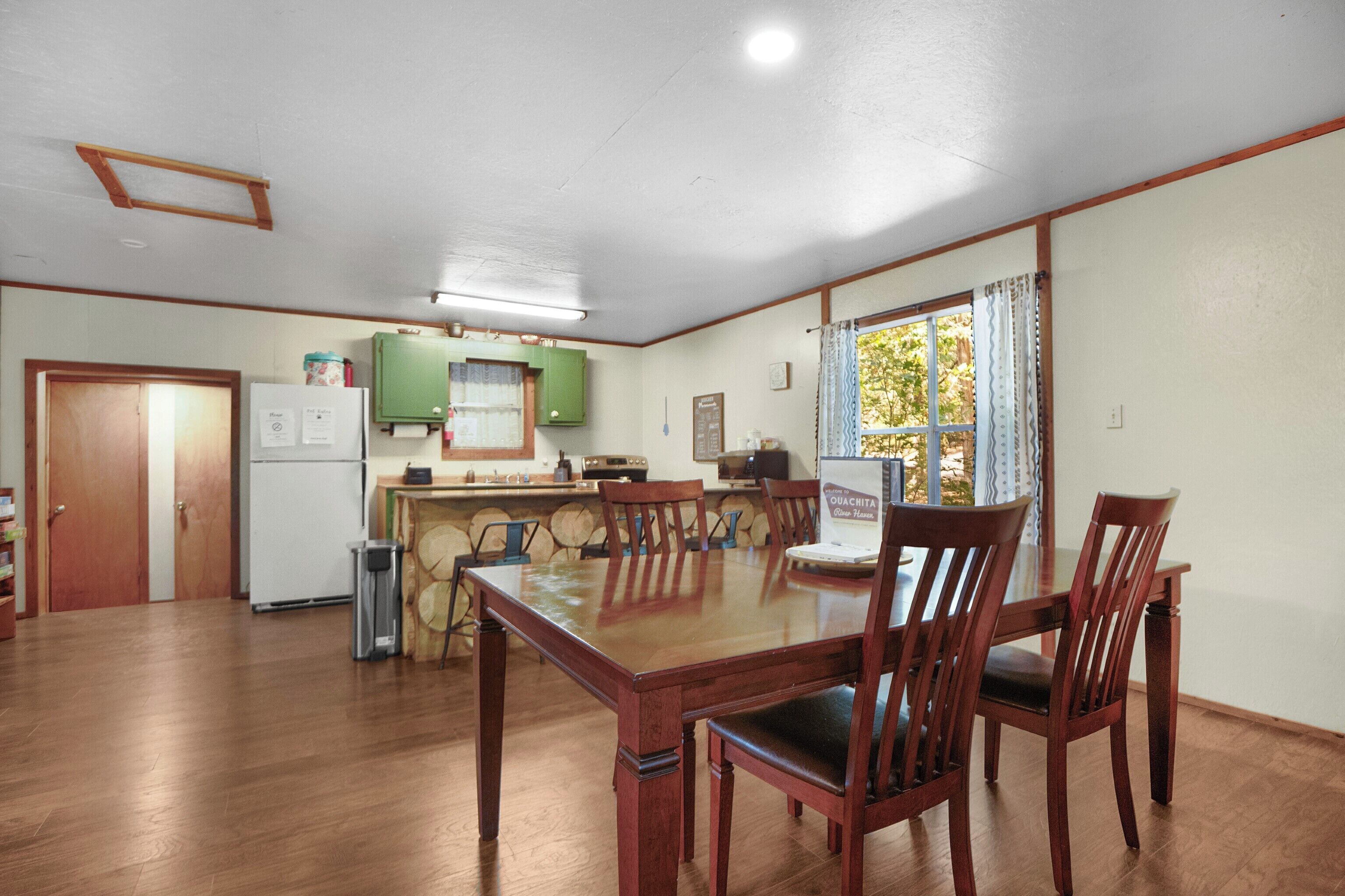 A dining area with a wooden table and chairs, a kitchen with green cabinets, and a window with curtains.