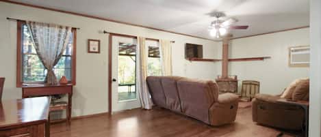 A spacious living room with a wooden floor, a sofa, a chair, a dining table, and a ceiling fan.