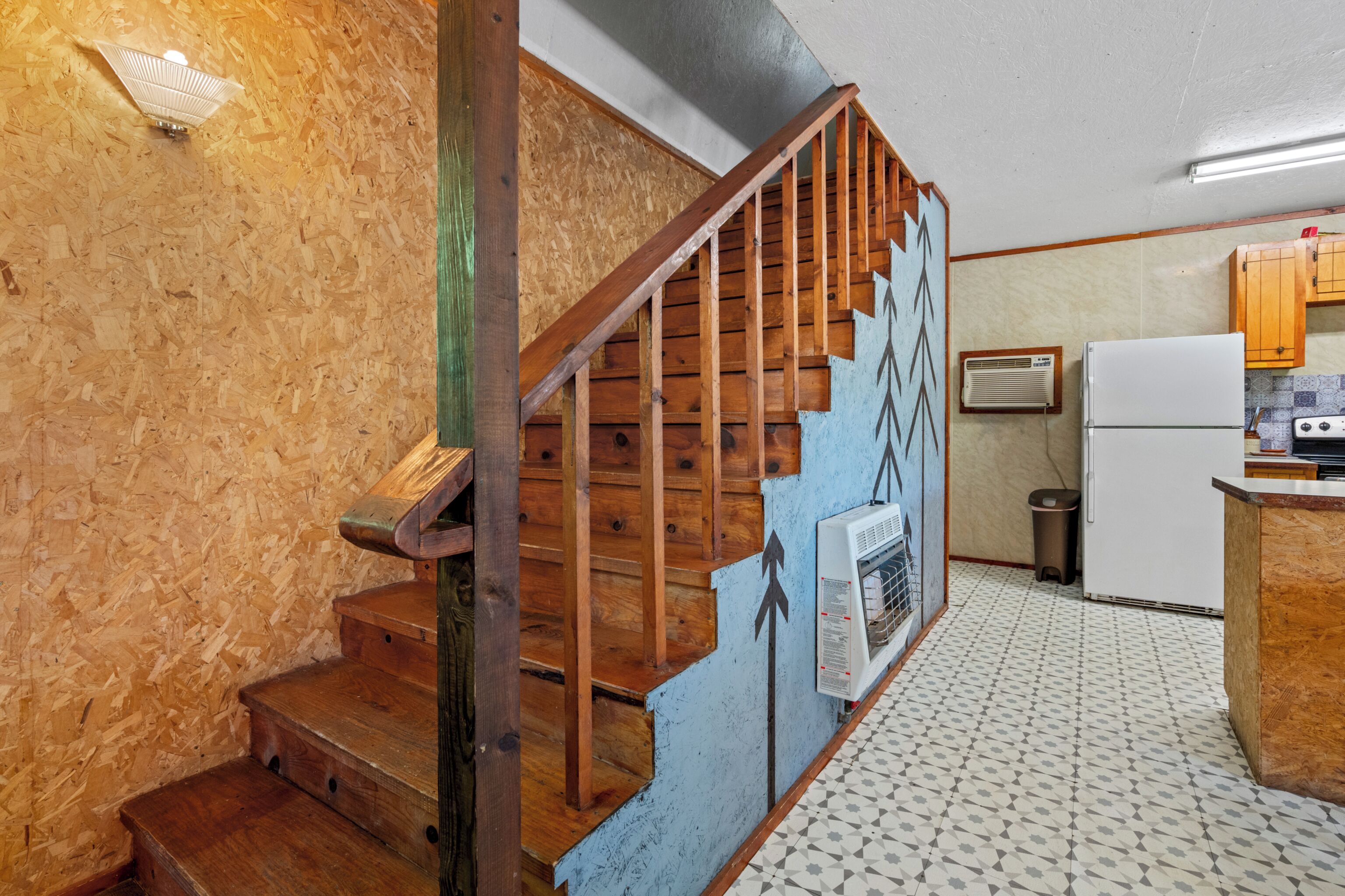 A wooden staircase with a handrail, a wall-mounted air conditioning unit, a white refrigerator, and a tiled floor.