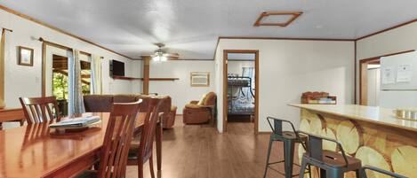 A spacious room with a wooden dining table, chairs, a bar area with stools, and a ceiling fan.