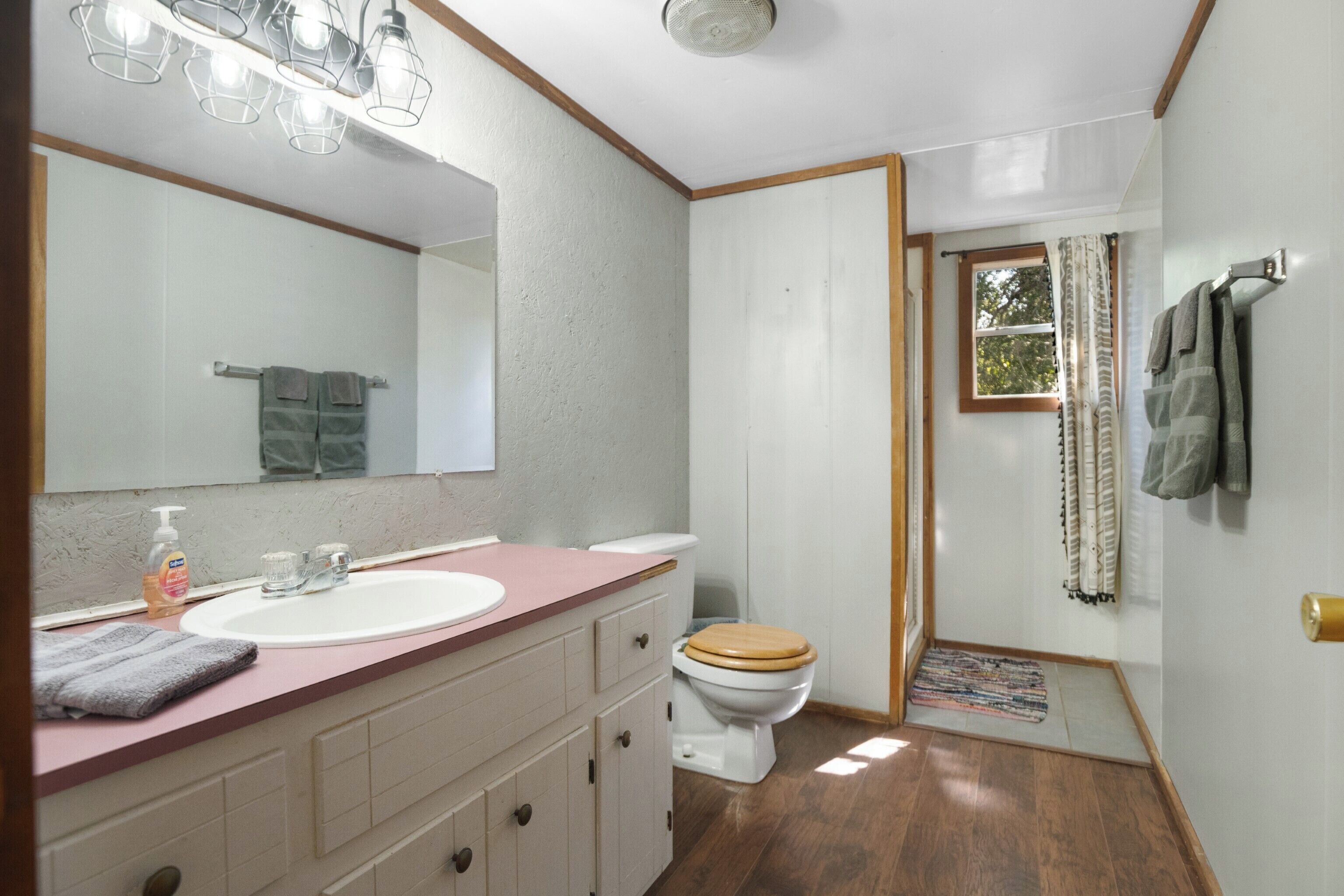 A bathroom with a wooden floor, a white sink, a wooden toilet, a mirror with lights, and a window with curtains.