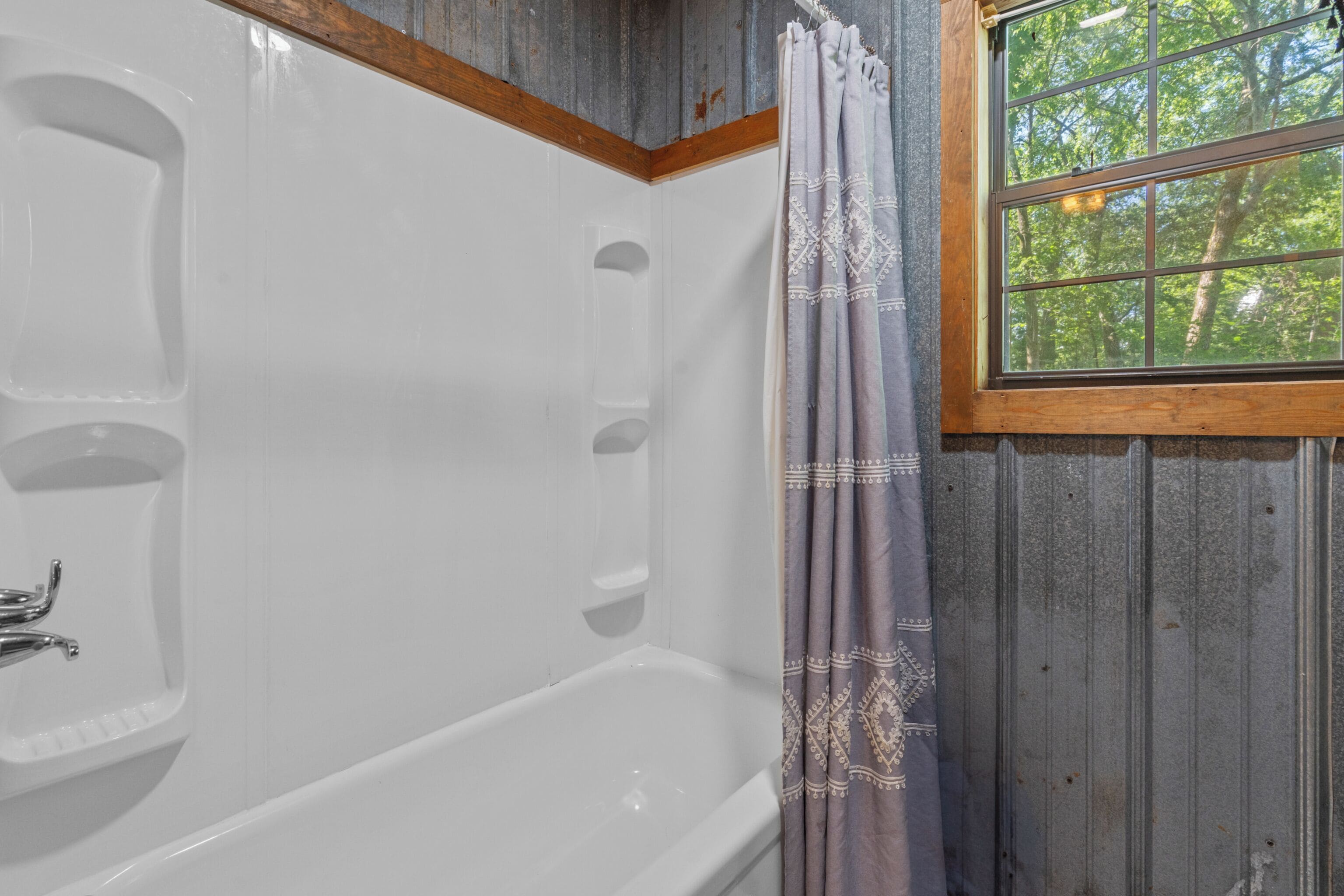 A bathroom with a bathtub, a window with a view of trees, and wooden walls.