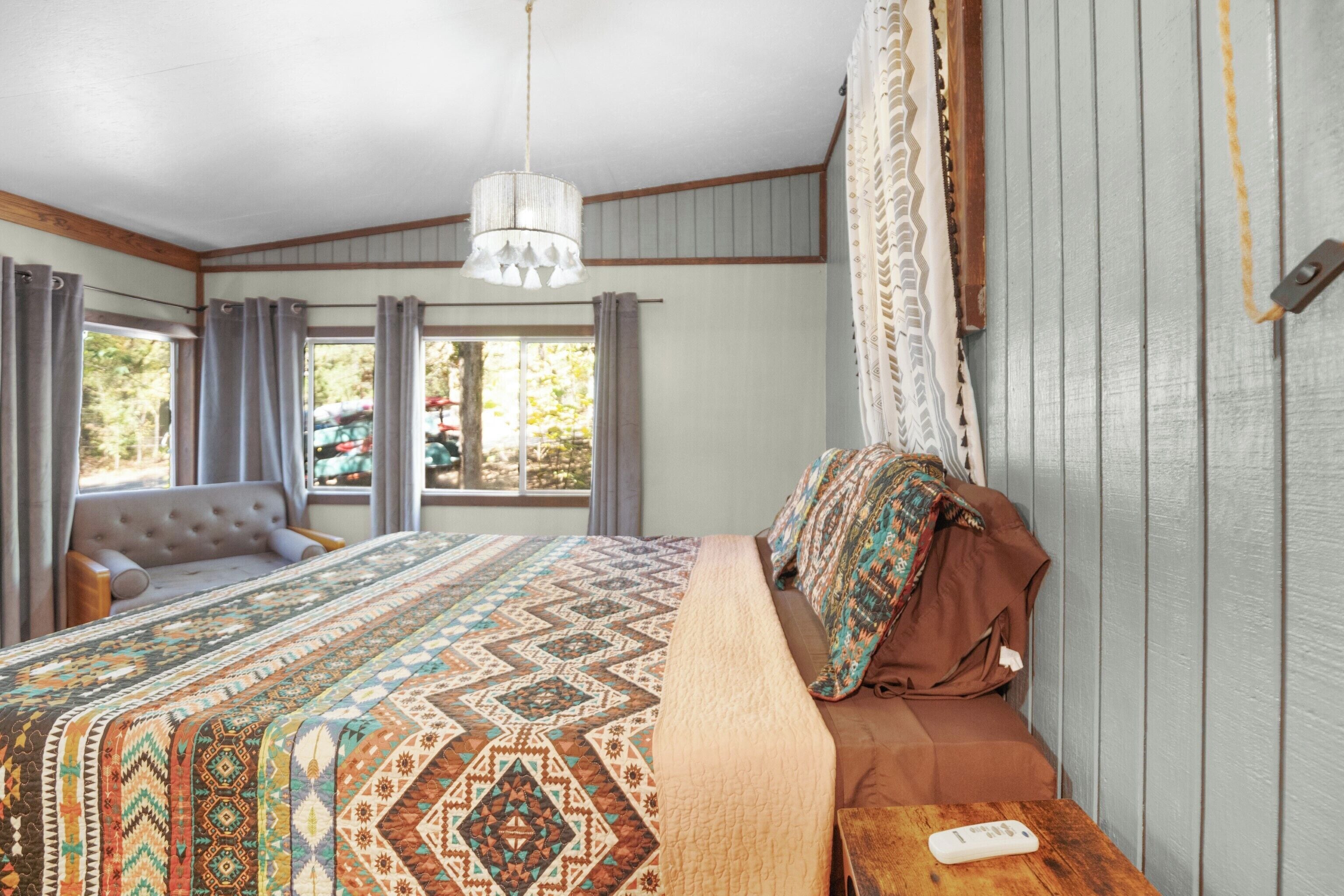Un dormitorio con una cama grande, una mesita de noche de madera, una lámpara colgante y un ventanal con cortinas.
