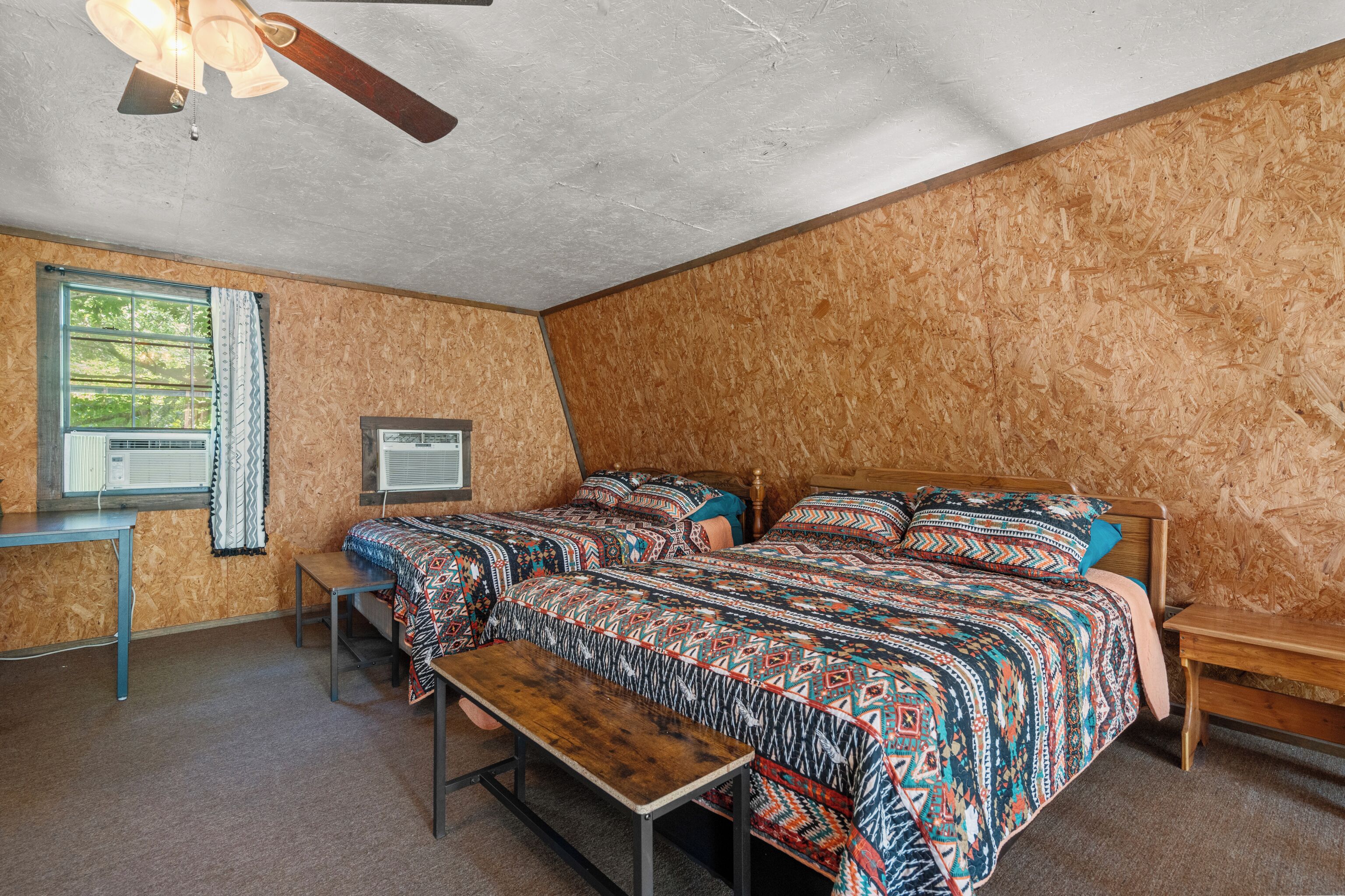 Ein Zimmer mit zwei Betten, einem Schreibtisch, einer Bank, einem Deckenventilator und einem Fenster mit Vorhängen.