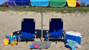 Un montaje en la playa con dos sillas azules, un paraguas colorido, un refrigerador, juguetes para la playa y toallas.