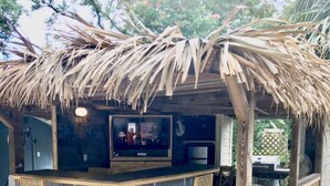 An outdoor bar area with a thatched roof, two wooden bar stools, and a television screen.