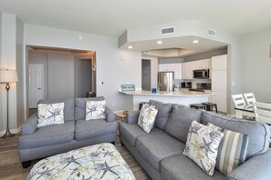 Een moderne woonkamer met grijze banken en kussens met zeesterrenmotief, een keuken met witte kasten en roestvrijstalen apparatuur, en een kleine eethoek met witte stoelen.