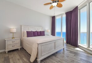 Een slaapkamer met een groot bed, nachtkastjes, een plafondventilator en uitzicht op de oceaan door grote ramen.