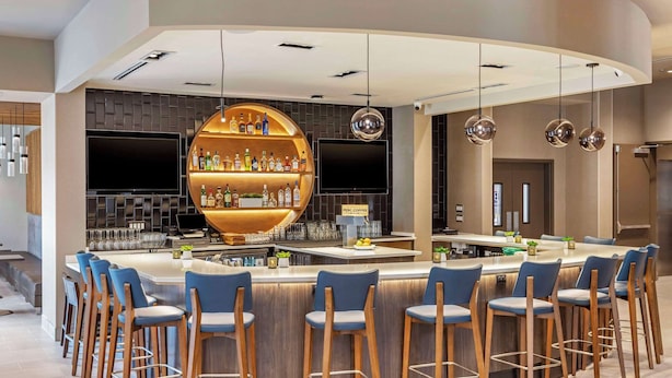 Zona de bar moderna con una estantería circular central que exhibe botellas, un televisor de pantalla plana y una barra con taburetes.