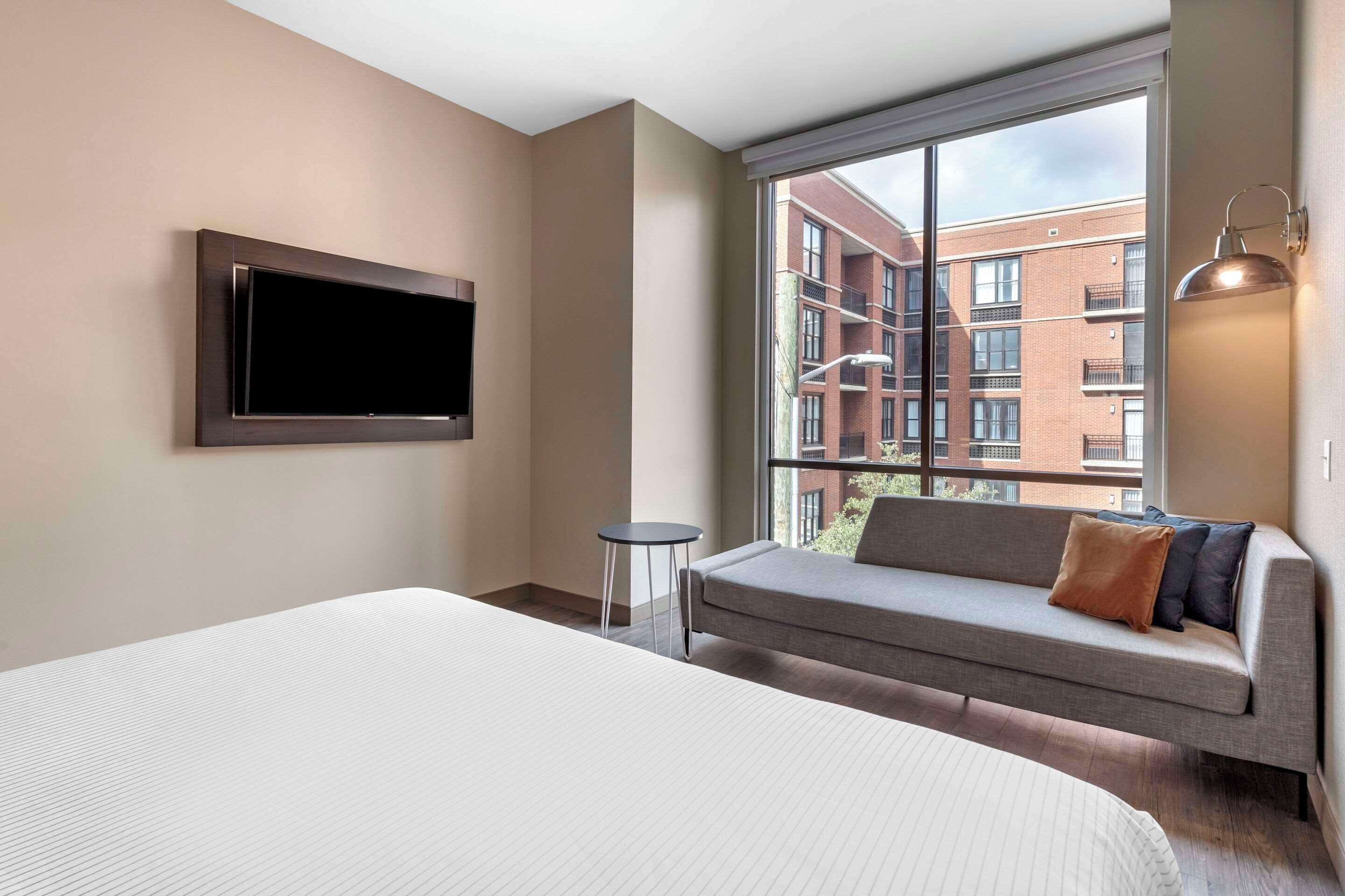 A modern hotel room with a large bed, a sofa with cushions, a TV mounted on the wall, a small table, and a large window offering a view of the building's exterior.