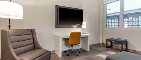 A hotel room with a large armchair, a desk with a computer, a television mounted on the wall, a window with a view, and a patterned rug.