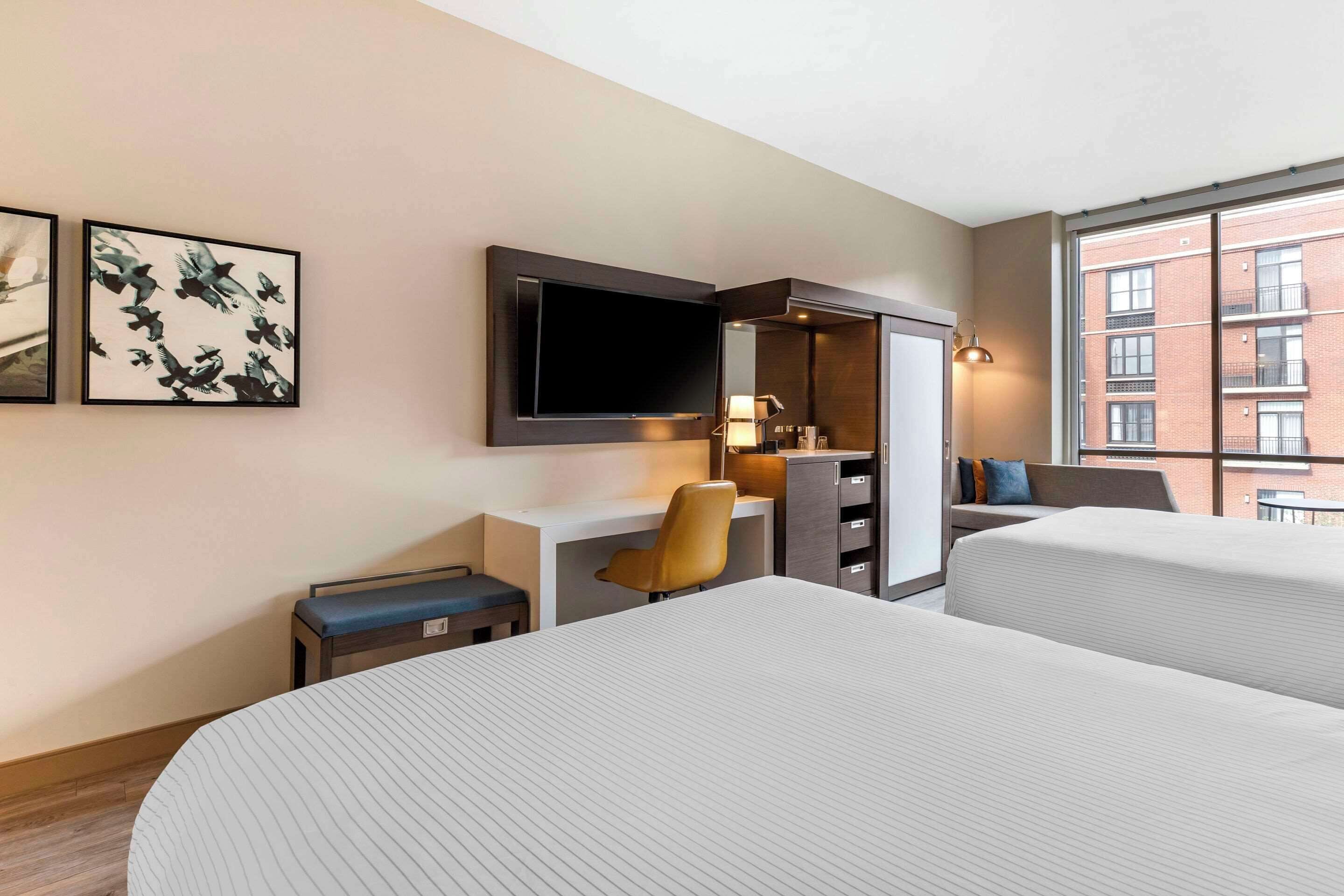 A hotel room with two beds, a desk, a chair, a TV, and a window with a view of buildings.