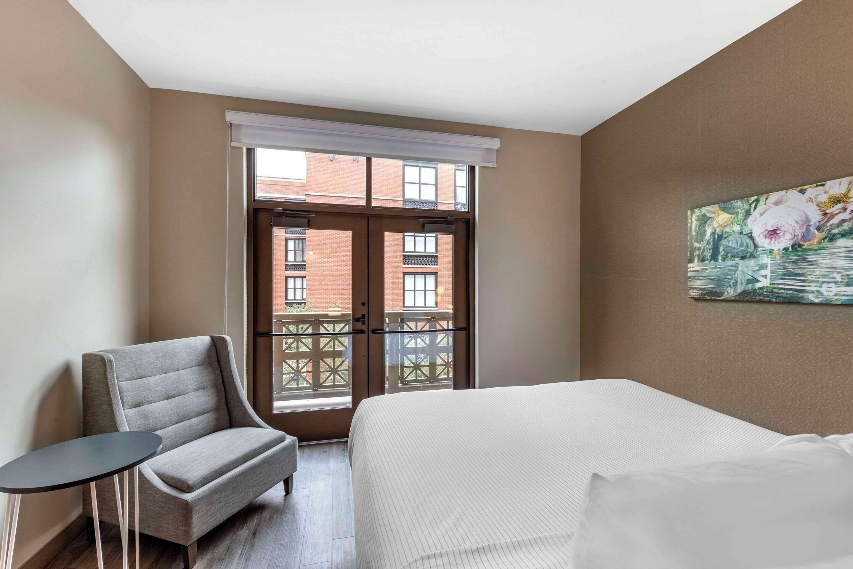 A hotel room with a bed, a chair, a small table, a window with a view of buildings, and a painting on the wall.