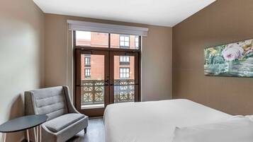 A hotel room with a bed, a chair, a small table, a window with a view of buildings, and a painting on the wall.
