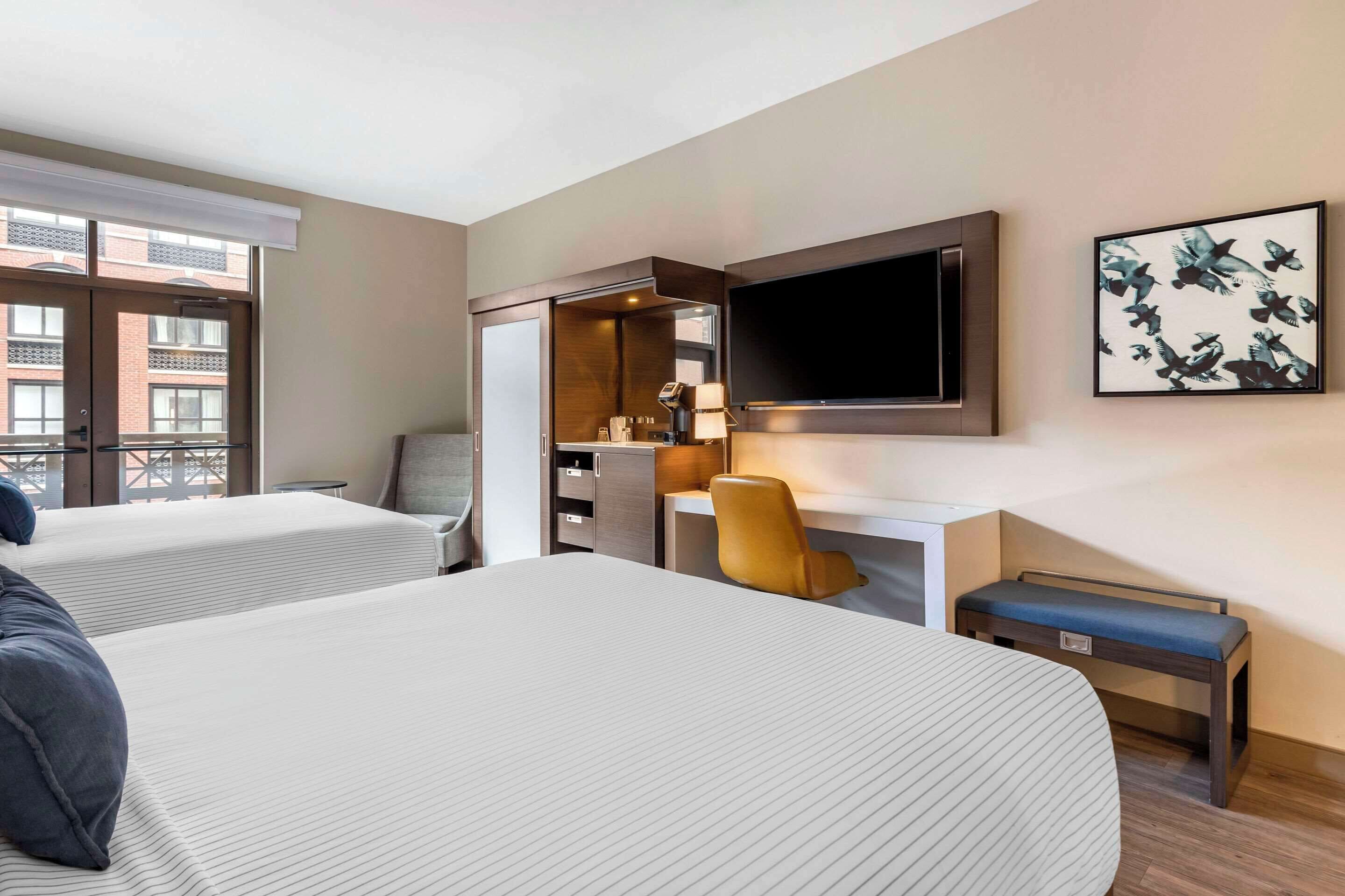 A hotel room with two beds, a desk, a chair, a TV, and a window with a view of buildings.