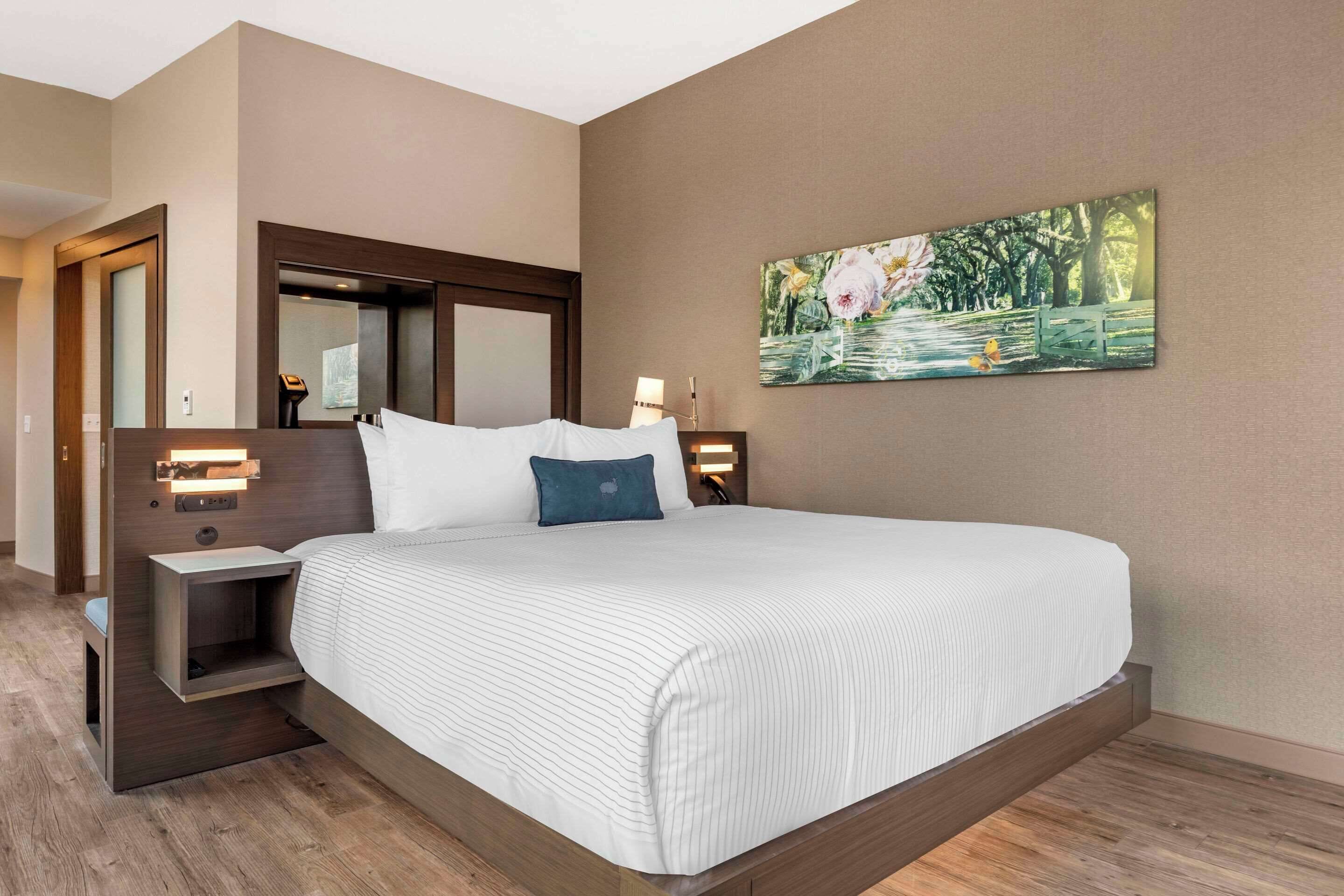 A hotel room with a large bed, a nightstand, a wall-mounted lamp, and a painting of a park scene.