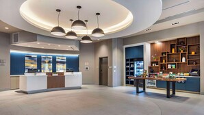 Un moderno vestíbulo de hotel con una recepción, una barra de café y diversas comodidades, incluyendo una estantería y una exhibición de aperitivos.
