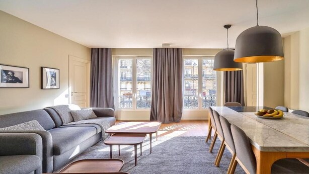 Un salon moderne avec un canapé, une table basse et une table à manger. On y trouve des rideaux, un téléviseur et une fenêtre donnant sur des immeubles.