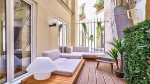Un espace de détente extérieur moderne, avec un canapé blanc, une petite table et une lampe. On y trouve également un pot de fleurs et une clôture en métal ornée de plantes grimpantes.