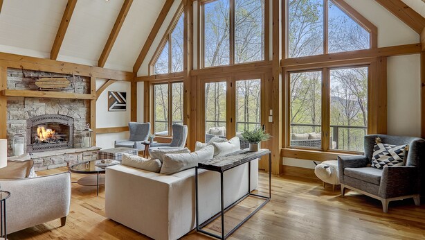 A spacious living room with a stone fireplace, large windows, and wooden beams.