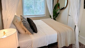 A bedroom with a bed, a bedside lamp, a window with blinds, a mirror, and a potted plant.