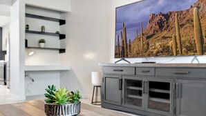 A modern living room with a flat-screen TV mounted on the wall, a wooden dining table with a potted plant, and a grey kitchen area with cabinets. - Casa Azul - Pool, ASU, Mill Ave, Sky Harbor, Old Town (Tempe)