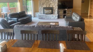 A spacious living room with a stone fireplace, leather sofas, a dining table with placemats, and a flat-screen TV mounted on the wall.