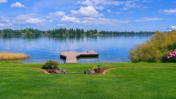 A serene lakeside view with a dock, grassy areas, and a distant mountain.