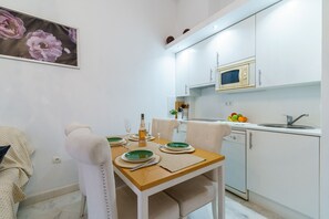 Uma cozinha compacta com armários brancos, micro-ondas e pia. Uma mesa de jantar com cadeiras acomoda dois lugares, e um quadro de flores está pendurado na parede.
