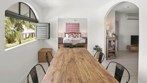 A dining area with a wooden table and chairs, a bedroom with a bed and a patterned wall hanging, and a bookshelf with books and a plant.