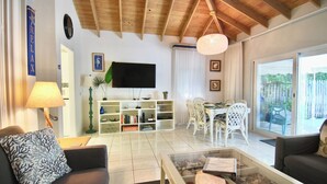 Una sala de estar con televisor, estanterías, zona de comedor y vistas amplias por grandes ventanales.