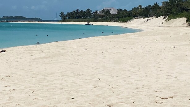Una extensa playa de arena clara con agua turquesa cristalina, palmeras y algunos edificios al fondo.