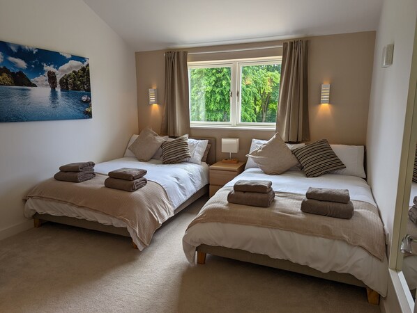 A bedroom with two beds, each with a pillow and a folded towel. There is a window with curtains, a nightstand with a lamp, and a painting on the wall. - Unique, Spacious, Self-Built Eco House with Huge Garden and Movie Room (Andover)