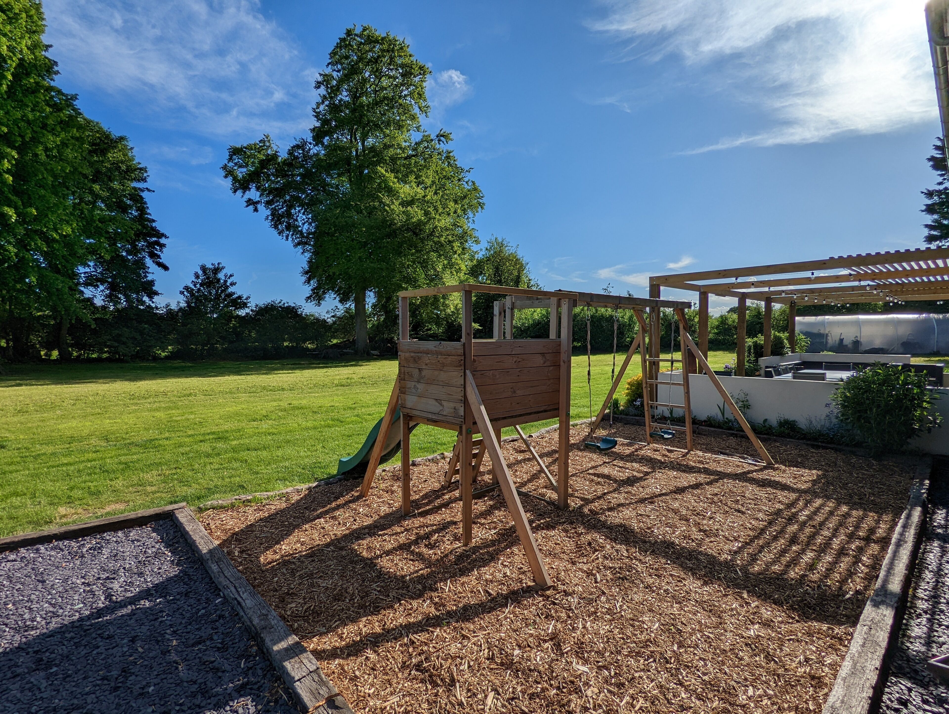 A wooden swing set in a grassy backyard with a pergola and outdoor seating area.
