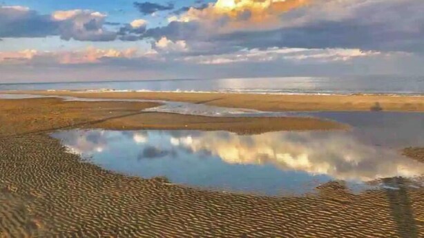 A beach with calm waters reflecting the sky and clouds, a sandy shore, and a distant horizon.