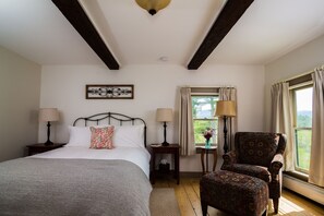 Um quarto com cama, duas mesas de cabeceira, uma cadeira, uma janela com cortinas e um objeto decorativo na parede.