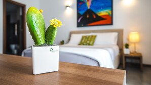 Una planta de cactus con una flor amarilla en una maceta blanca sobre una mesa de madera, con una cama y un cuadro colorido al fondo.
