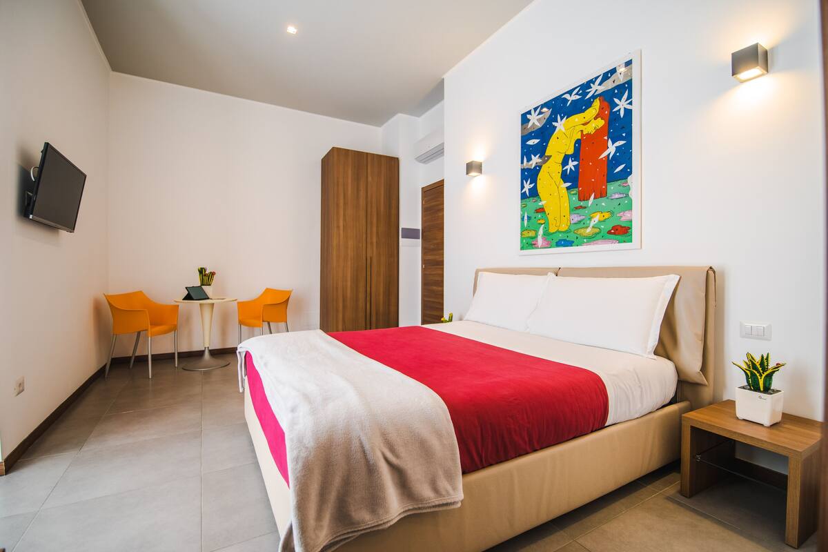 A hotel room with a bed, a small table with chairs, a TV mounted on the wall, and a painting above the bed.