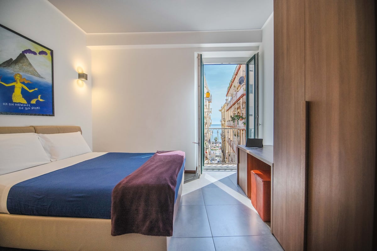 A hotel room with a bed, a desk, a view of the street, and a painting on the wall.