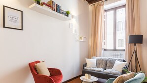 A living room with a red chair, a teal armchair, a sofa, a floor lamp, and a bookshelf with books and a plant. - Serpenti Apartment (Roma)