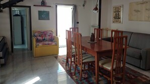 Ein gemütlicher Wohnbereich mit einem Holzesstisch, Stühlen, einer Couch und einem bunten Teppich. Im Hintergrund ist eine Treppe mit Holzbalken zu sehen.