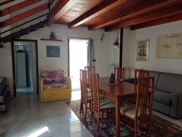 A cozy living area with a wooden dining table, chairs, a sofa, and a colorful rug. A staircase with wooden beams is visible in the background.