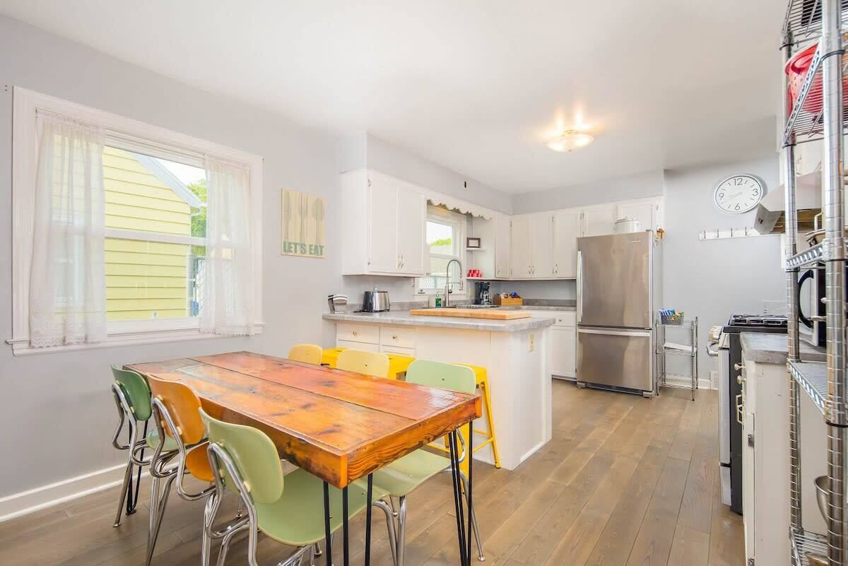 A modern kitchen with white cabinets, a stainless steel refrigerator, and a wooden dining table with colorful chairs.