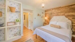 Un dormitorio con cabecera de madera, colcha blanca y mesita de noche con lámpara. Se observa un armario blanco con estantes y una cesta en la parte superior.