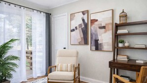 A room with a large window, a chair, a desk, and two framed abstract paintings.