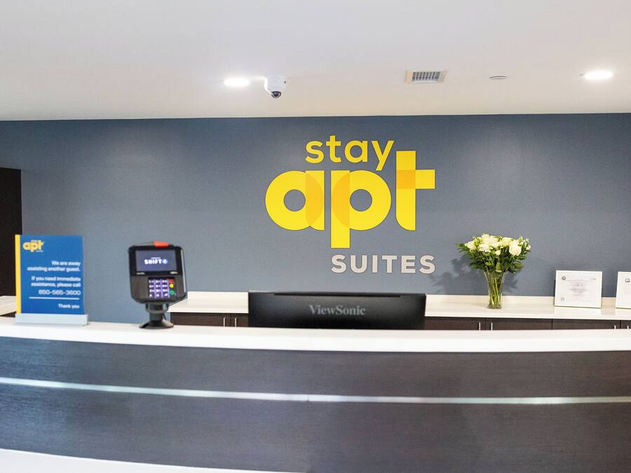 A reception desk with a 'stay apt SUITES' sign, a ViewSonic monitor, a payment terminal, and a vase with flowers.