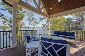 Una terraza techada con muebles blancos y cojines azules, con vista a un lago y una casa al fondo.