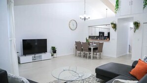 A modern living room with a flat-screen TV, a glass coffee table, and a dining area with a white table and chairs.