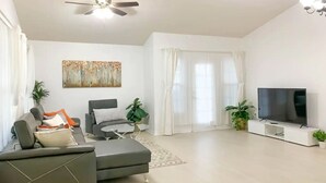 A modern living room with a grey sofa, a small round table, a ceiling fan, a flat-screen TV, and a large window with white curtains.